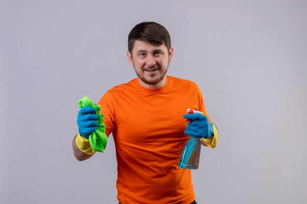 Joven vestido con camiseta naranja y guantes de goma con spray de limpieza y alfombra sonriendo alegremente