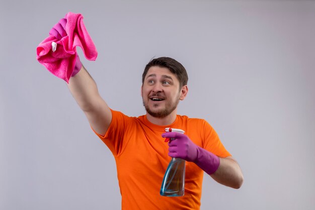 Joven vestido con camiseta naranja y guantes de goma con spray de limpieza y alfombra sonriendo alegremente