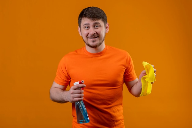 Joven vestido con camiseta naranja y guantes de goma con spray de limpieza y alfombra sonriendo alegremente