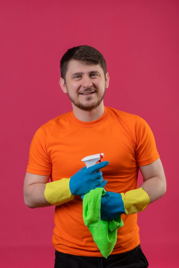 Foto gratuito joven vestido con camiseta naranja y guantes de goma con spray de limpieza y alfombra sonriendo alegremente
