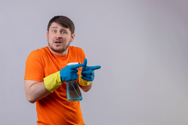 Joven vestido con camiseta naranja y guantes de goma con spray de limpieza preocupado apuntando con los dedos hacia el lado