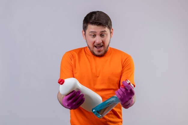 Joven vestido con camiseta naranja y guantes de goma con suministros de limpieza mirándolos