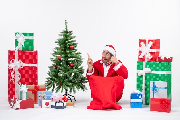 Foto gratuita joven vestido como papá noel con regalos y árbol de navidad decorado sentado en el suelo haciendo gesto de llamarme y apuntando arriba