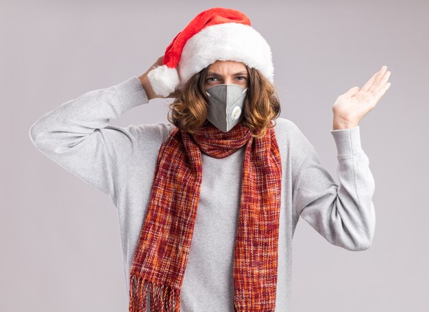 Joven vestido con gorro de navidad de santa y máscara protectora facial con bufanda caliente alrededor de su cuello confundido y disgustado de pie sobre la pared blanca