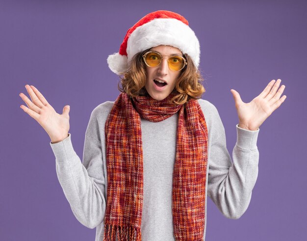 Joven vestido con gorro de Papá Noel de Navidad y gafas amarillas con bufanda alrededor de su cuello mirando a la cámara sorprendido y feliz con los brazos levantados sobre fondo púrpura