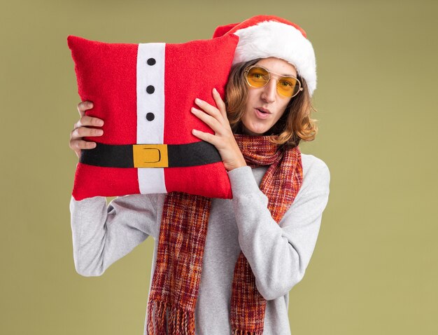 Joven vestido con gorro de Papá Noel de Navidad y gafas amarillas con cálida bufanda alrededor de su cuello sosteniendo una almohada de Navidad mirando a la cámara con una sonrisa en la cara de pie sobre fondo verde