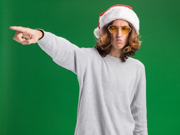 Joven vestido con gorro de Papá Noel de Navidad y gafas amarillas mirando a un lado con el ceño fruncido apuntando con el dedo índice al lado de pie sobre fondo verde