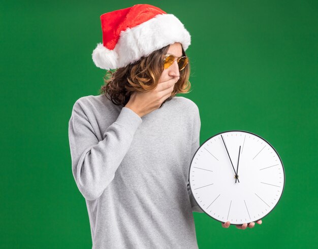 Joven vestido con gorro de Papá Noel de Navidad y gafas amarillas sosteniendo el reloj de pared mirándolo asombrado cubriendo la boca con la mano sobre la pared verde