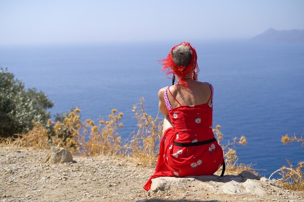 Una joven con un vestido rojo se sienta de espaldas a la cámara con una hermosa vista del mar desde la montaña disfruta de la vista y las vacaciones