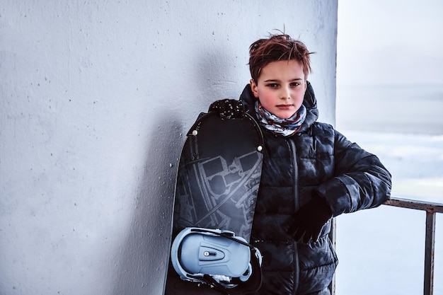 Foto gratuita un joven vestido con traje de nieve sosteniendo una tabla de snowboard apoyado en una baranda cerca de la playa nevada