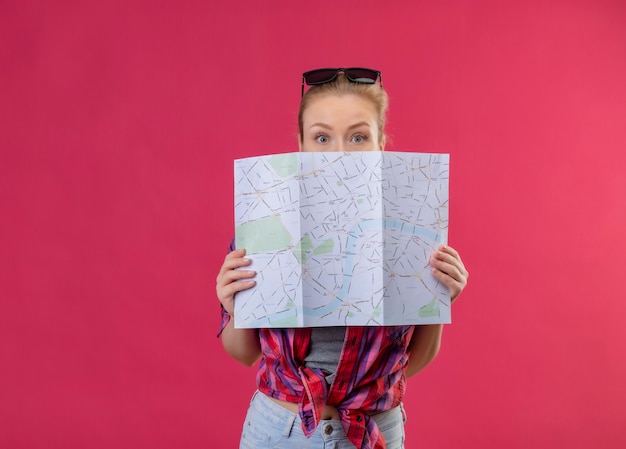 Foto gratuita joven viajera con camisa roja y gafas en la cabeza cubrió la boca con el mapa en la pared rosa aislada
