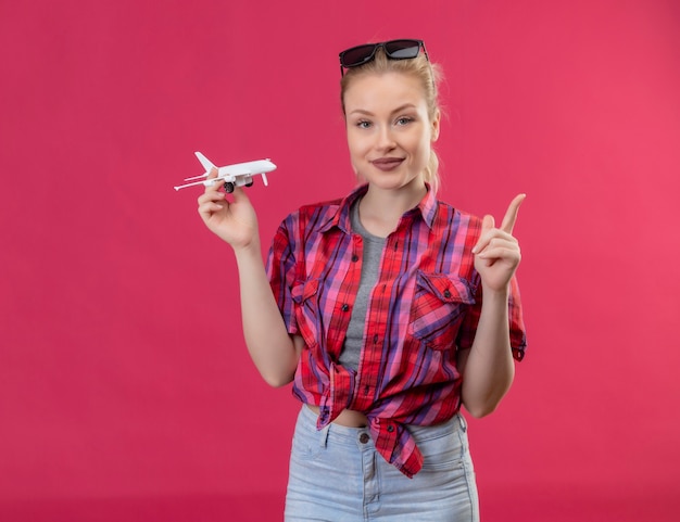 Foto gratuita joven viajera con camisa roja y gafas en la cabeza sosteniendo el plan de juguete apunta al lado de la pared rosa aislada