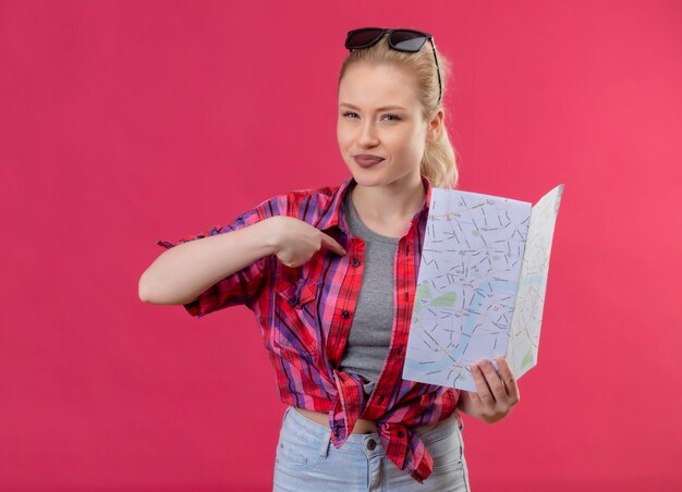 Joven viajera vestida con camisa roja y gafas en la cabeza sosteniendo el mapa apunta a sí misma en la pared rosa aislada