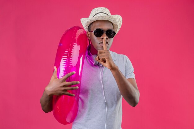 Joven viajero afroamericano hombre con sombrero de verano con auriculares alrededor de su cuello con gafas de sol negras sosteniendo anillo inflable haciendo gesto de silencio con el dedo en liips sobre pi