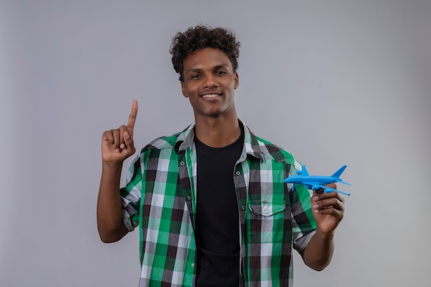 Joven viajero afroamericano hombre sosteniendo avión de juguete apuntando con el dedo hacia arriba sonriendo con expresión de confianza en la cara de pie sobre fondo blanco.