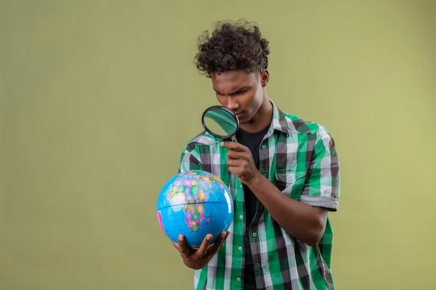 Joven viajero afroamericano hombre sujetando el globo mirando a través de una lupa con interés de pie sobre fondo verde