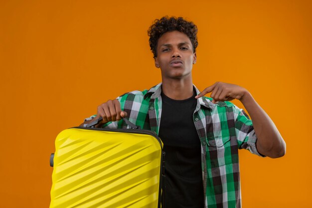 Joven viajero afroamericano hombre sujetando la maleta apuntando con el dedo a sí mismo mirando seguro de sí mismo satisfecho sobre fondo naranja