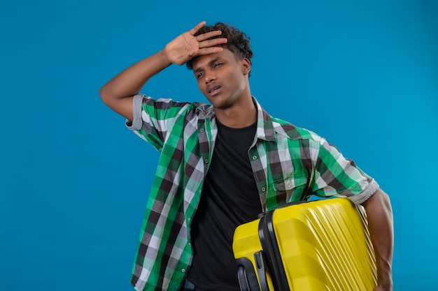 Joven viajero afroamericano hombre sujetando la maleta con aspecto cansado y aburrido de pie sobre fondo azul.