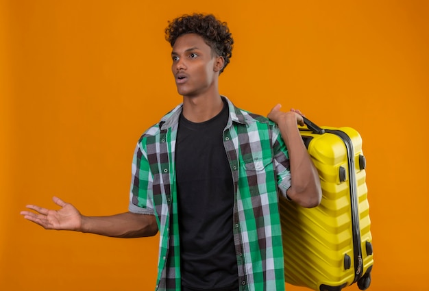 Joven viajero afroamericano hombre sujetando la maleta mirando a un lado confundido sin tener respuesta de pie sobre fondo naranja