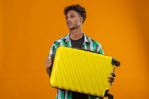 Joven viajero afroamericano hombre sujetando la maleta mirando a un lado con seria expresión de confianza en la cara de pie sobre fondo naranja