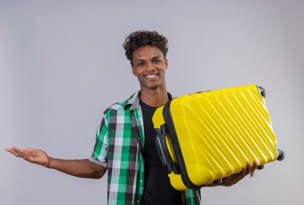 Joven viajero afroamericano hombre sujetando la maleta presentando copia espacio con el brazo de su mano sonriendo alegremente de pie sobre fondo blanco.