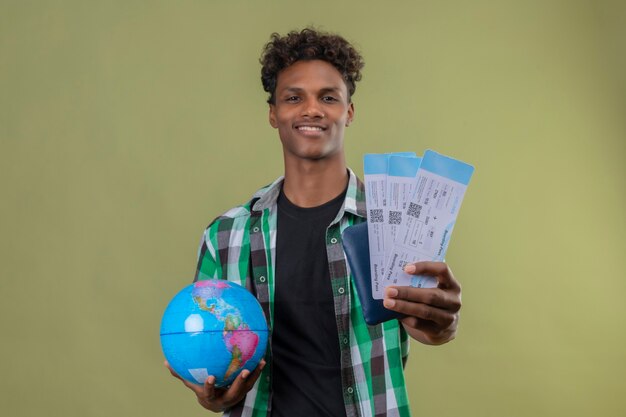 Joven viajero afroamericano hombre sujetando el mundo mostrando billetes de avión sonriendo alegremente de pie sobre fondo verde