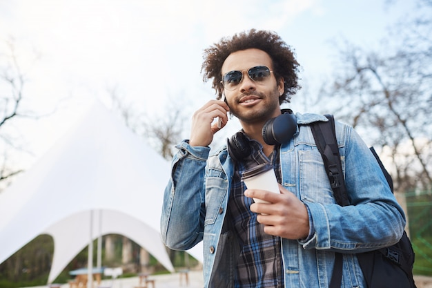 Joven viajero afroamericano con peinado afro sosteniendo café mientras camina y habla por teléfono inteligente, mirando a un lado con expresión concentrada y complacida.