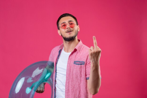 Joven viajero guapo chico con gafas de sol con anillo inflable mirando a la cámara alegre mostrando el dedo medio parado sobre fondo rosa