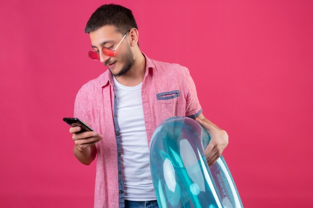 Joven viajero guapo chico con gafas de sol con anillo inflable mirando la pantalla de su móvil sonriendo seguro de pie sobre fondo rosa