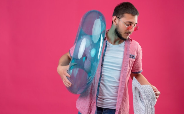 Joven viajero guapo chico con gafas de sol con anillo inflable y sombrero de verano mirando a un lado con expresión seria y segura en la cara de pie sobre fondo rosa