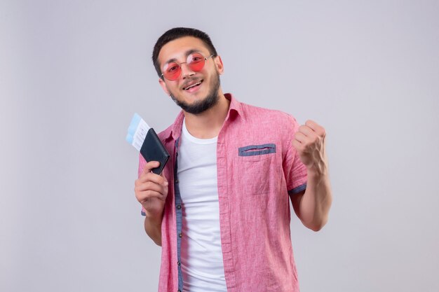 Joven viajero guapo chico con gafas de sol con boletos aéreos mirando a la cámara salió y feliz levantando el puño después de un concepto ganador de la victoria de pie sobre fondo blanco