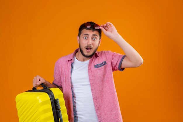 Joven viajero guapo chico sosteniendo la maleta tocando sus gafas de sol en la cabeza mirando a la cámara sorprendido y salido de pie sobre fondo naranja
