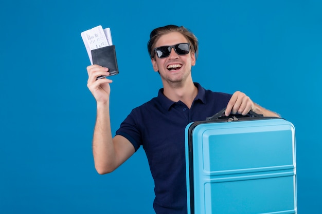 Joven viajero guapo hombre que llevaba gafas de sol negras de pie con la maleta con billetes de avión mirando a la cámara con cara feliz sonriendo alegremente sobre fondo azul