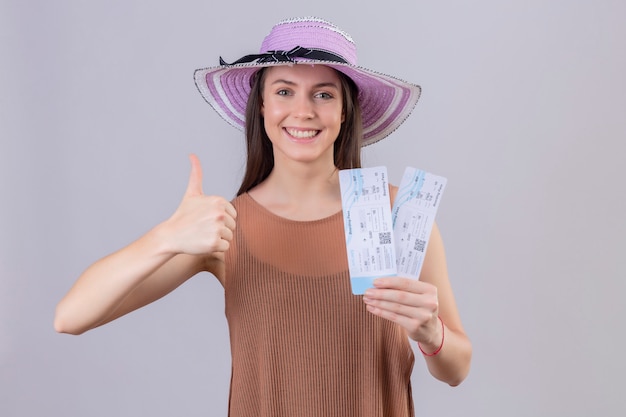 Joven viajero hermosa mujer con sombrero de verano con boletos aéreos sonriendo con cara feliz mostrando los pulgares para arriba sobre la pared blanca