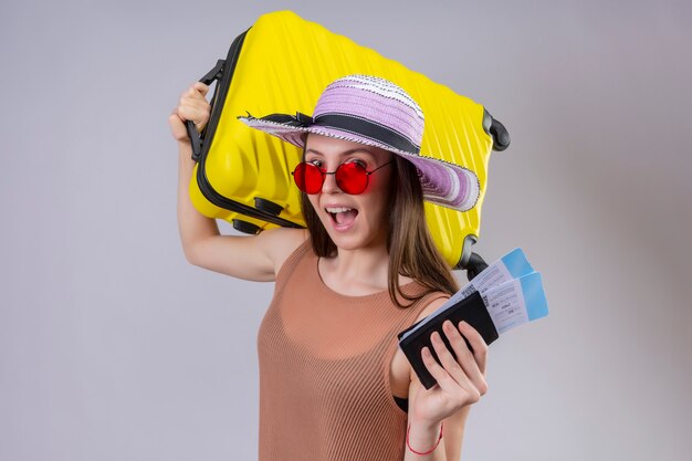 Joven viajero hermosa mujer con sombrero de verano con gafas de sol rojas con maleta amarilla y boletos aéreos sonriendo alegremente con cara feliz sobre pared blanca