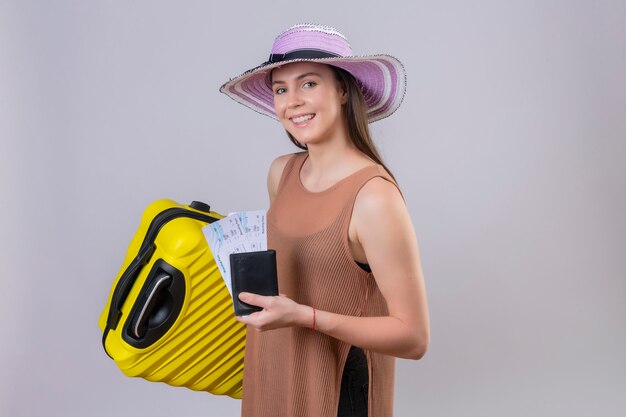 Joven viajero hermosa mujer con sombrero de verano con maleta amarilla y entradas sonriendo con cara feliz sobre pared blanca