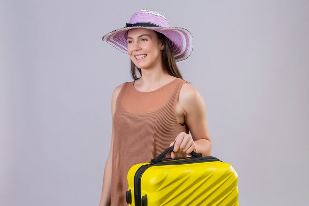 Joven viajero hermosa mujer con sombrero de verano con maleta amarilla sonriendo con cara feliz sobre pared blanca