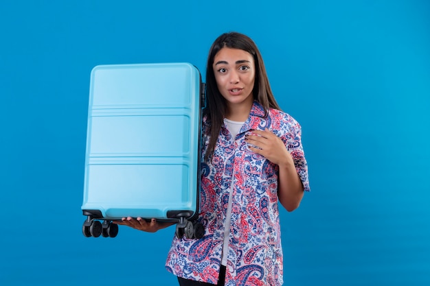 Foto gratuita joven viajero hermosa mujer sosteniendo la maleta azul mirando confundido y sorprendido sobre la pared azul