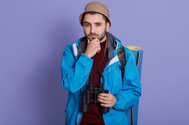 Joven viajero hombre caucásico confundido, se siente dudoso e inseguro, posando contra la pared azul con mochila y binoculares