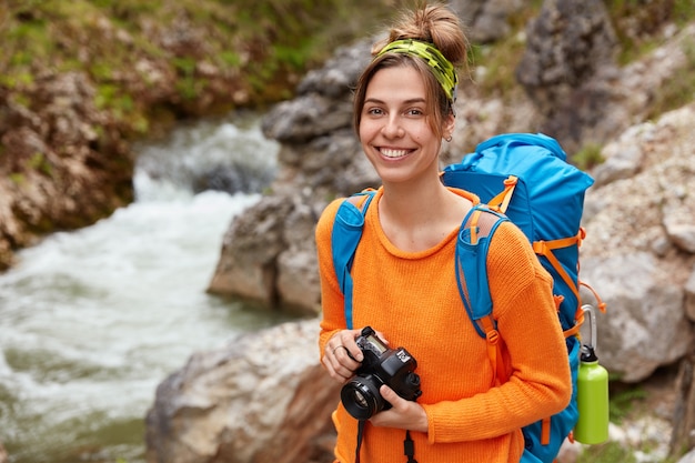 Joven viajero positivo posa con cámara y mochila contra el cañón, disfruta de la naturaleza y el paisaje