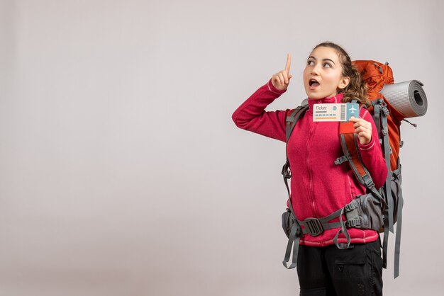 Joven viajero sorprendido con mochila grande sosteniendo el billete de viaje apuntando con el dedo hacia arriba