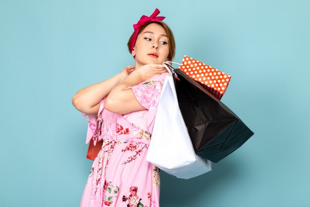 Una joven de vista frontal en vestido rosa diseñado con flores posando sosteniendo paquetes de compras en azul