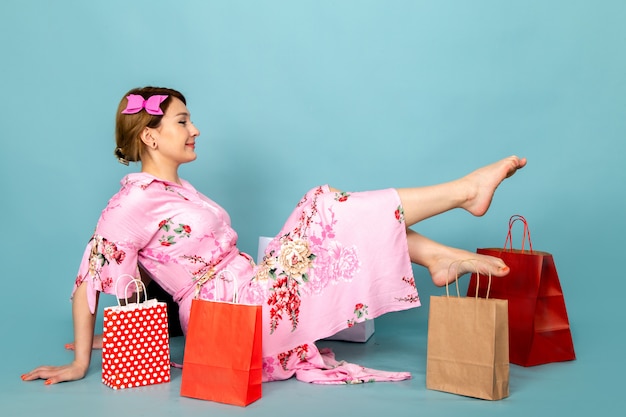 Una joven de vista frontal con vestido rosa diseñado con flores sentado y posando con una sonrisa y paquetes de compras en azul