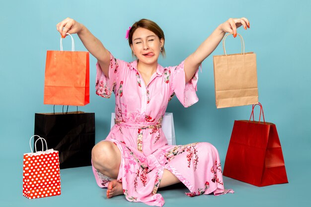 Una joven de vista frontal con vestido rosa diseñado con flores sentado y posando con una sonrisa y paquetes de compras en azul