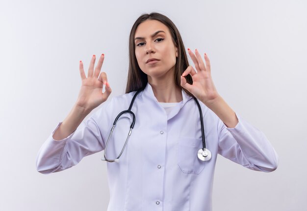 Joven vistiendo bata médica estetoscopio mostrando gesto okey en pared blanca aislada