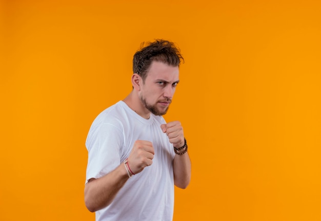 Joven vistiendo camiseta blanca de pie en pose de lucha en la pared naranja aislada