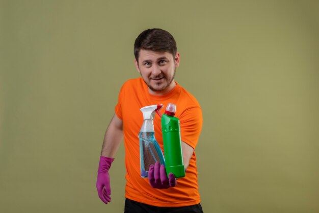 Joven vistiendo camiseta naranja y guantes de goma sosteniendo suministros de limpieza sonriendo feliz y positivo de pie sobre la pared verde 3