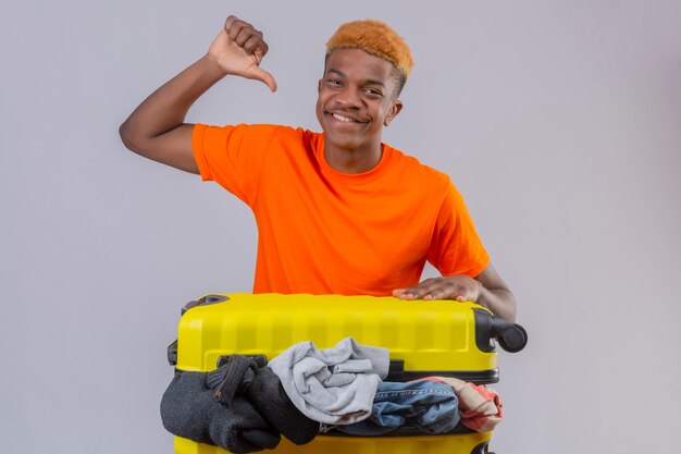 Joven vistiendo camiseta naranja de pie con maleta de viaje llena de ropa optimista y alegre sonriendo apuntando a sí mismo con el pulgar sobre la pared blanca