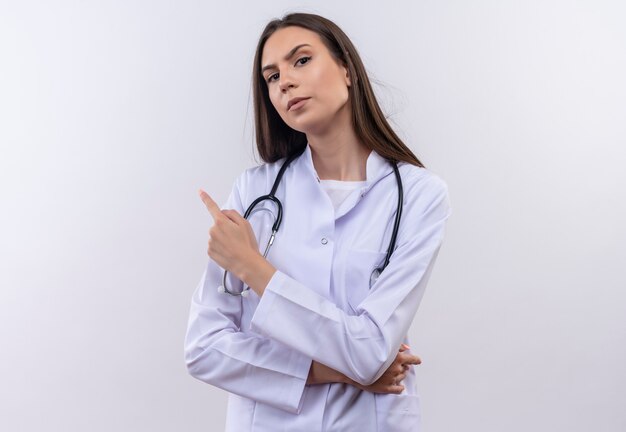 Joven vistiendo estetoscopio bata médica apunta al lado de la pared blanca aislada