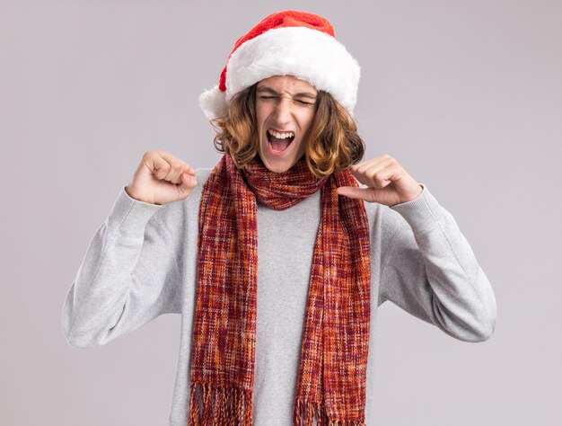 Joven vistiendo gorro de navidad de santa con bufanda caliente alrededor de su cuello apretando los puños loco feliz y emocionado de pie sobre la pared blanca